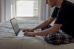 young-man-sitting-on-bed-working-on-laptop-in-morn